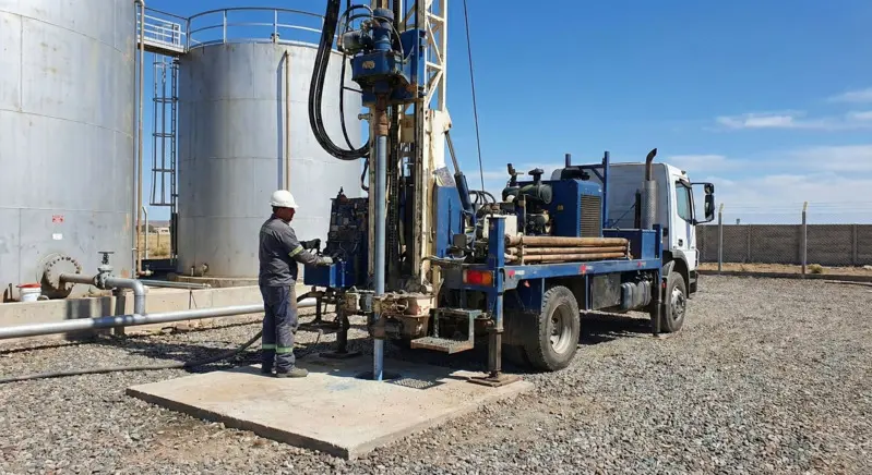 Equipo de perforación rotativo de gran capacidad operando en un predio industrial de Neuquén, realizando un pozo de monitoreo ambiental cerca de tanques de almacenamiento, bajo estrictas normas de seguridad.